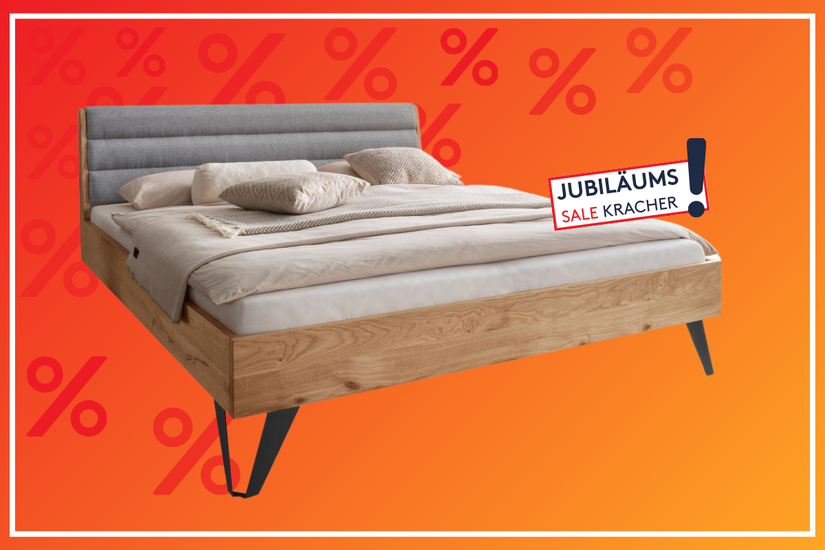 Holzbett mit gepolstertem, grauem Kopfteil und beigefarbener Bettwäsche auf orange-rotem Hintergrund mit Prozentzeichen und dem Hinweis „Jubiläums-Sale Kracher“.